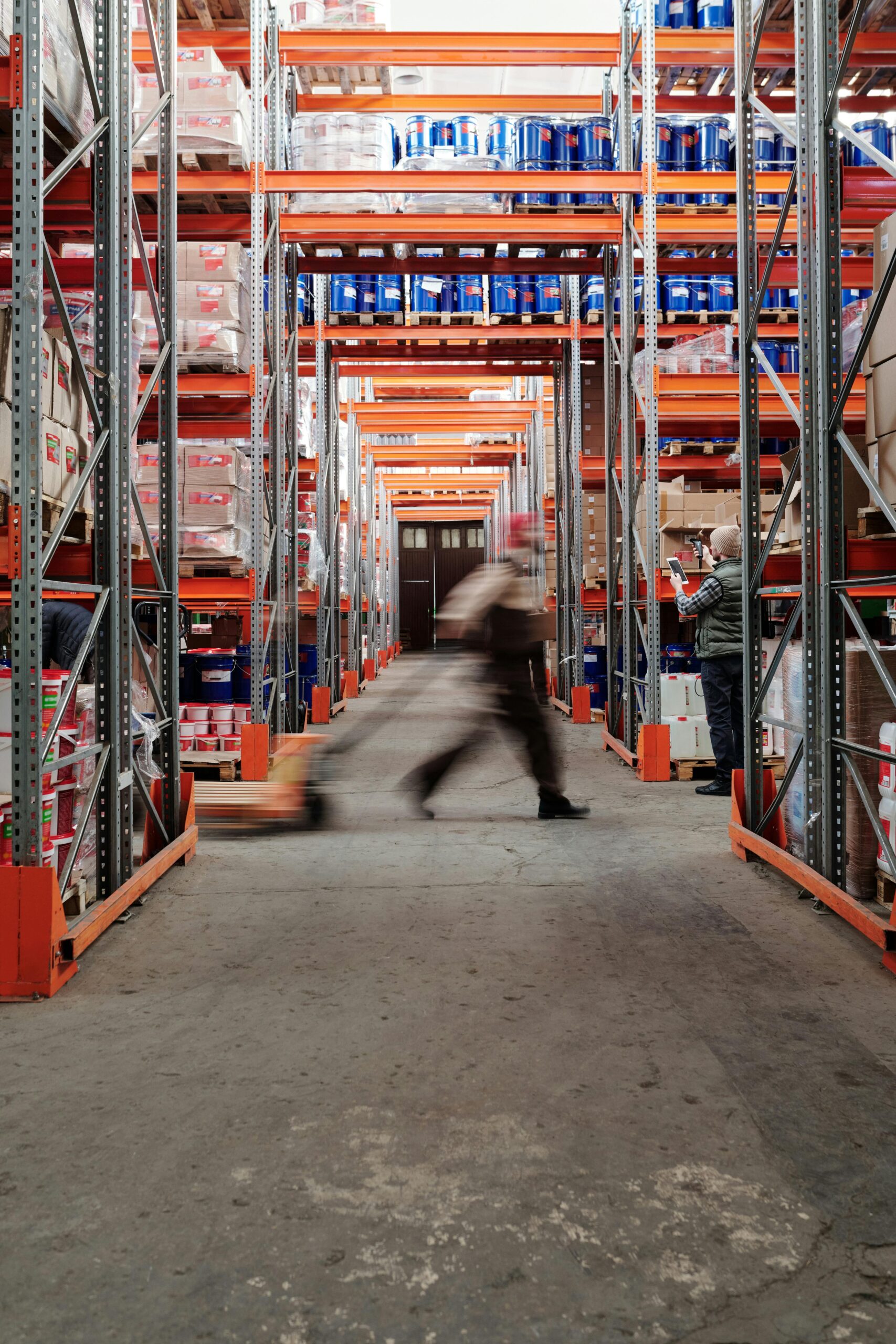 Blurred motion of a man walking in a spacious warehouse aisle between tall shelves.